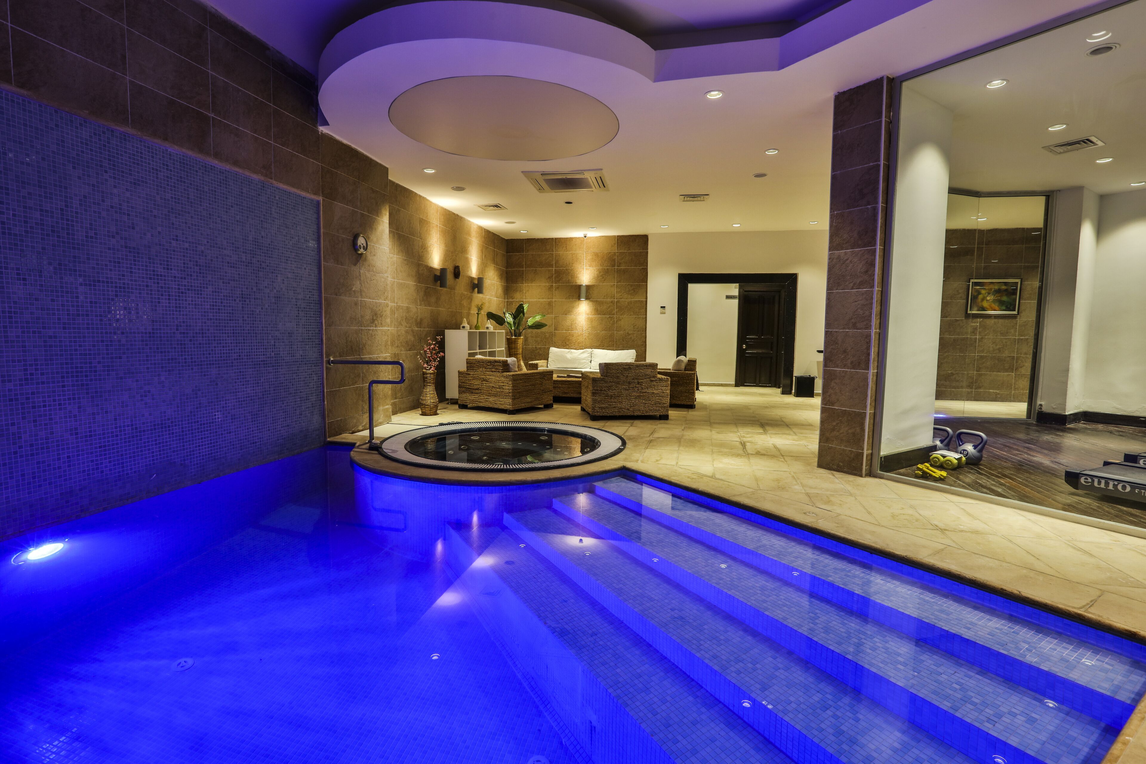 Un área de piscina interior moderna con un elemento acuático iluminado en azul, un espacio de descanso con sofá y sillones, y una recepción.