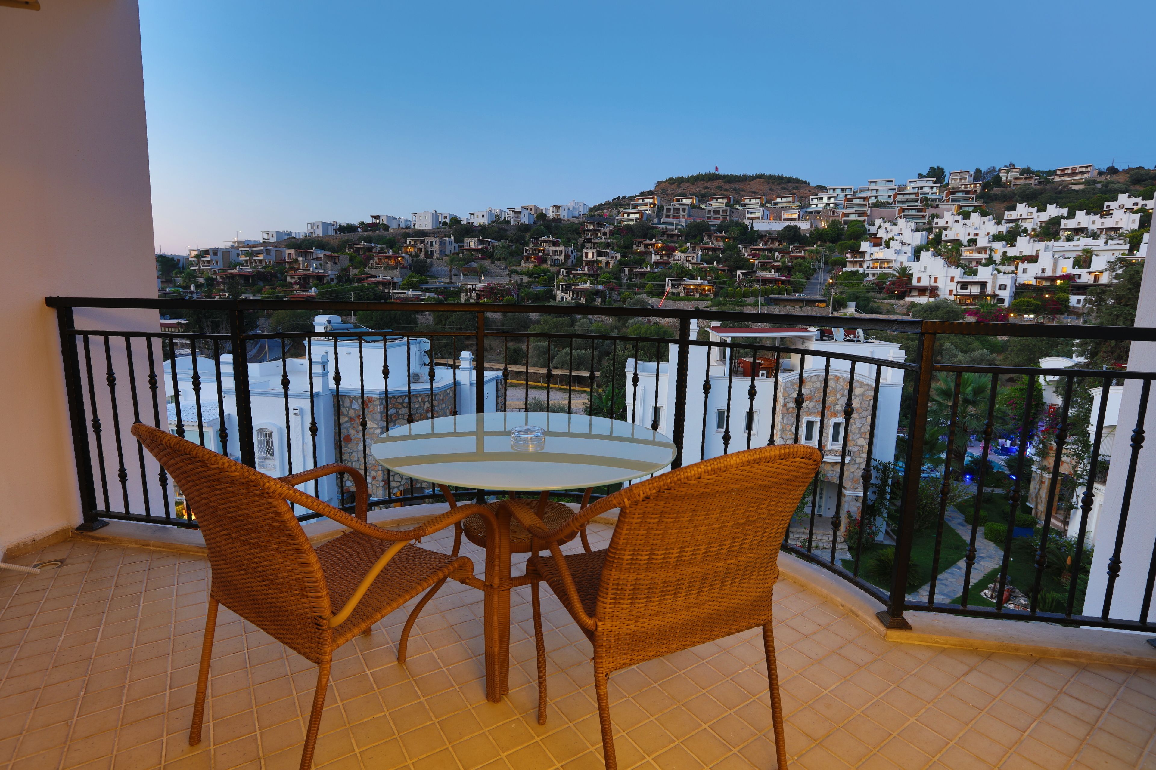 A balcony with wicker chairs and a table, overlooking a hillside of white buildings.