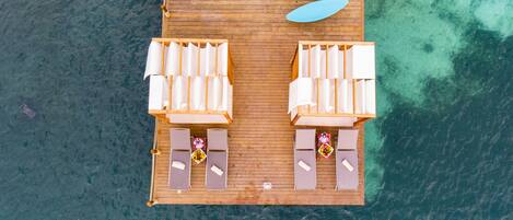 An aerial view of a wooden deck with lounge chairs and a surfboard, overlooking clear turquoise water.