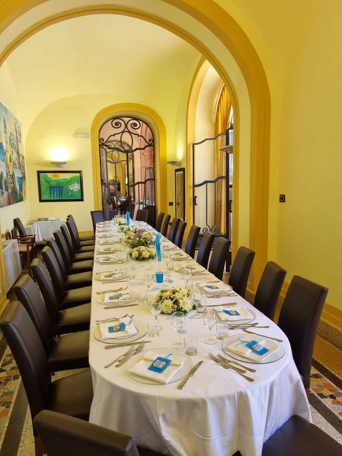 Uma longa mesa de jantar posta para uma refeição formal, rodeada por cadeiras, em um ambiente com paredes amarelas e uma porta em arco.