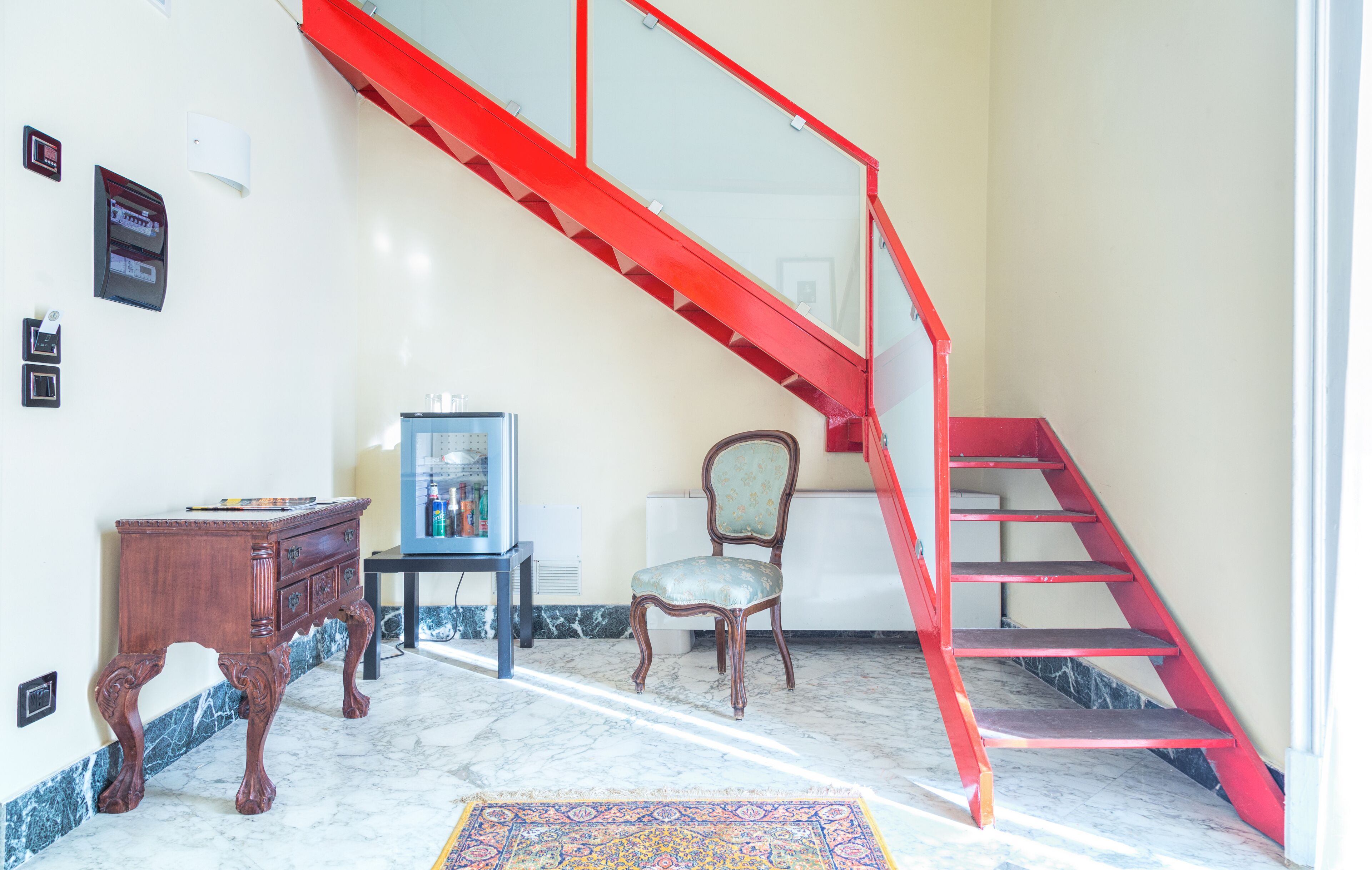 A staircase with red railings leading to an upper floor, a wooden desk with drawers, a chair, and a small table with a blue object on it.