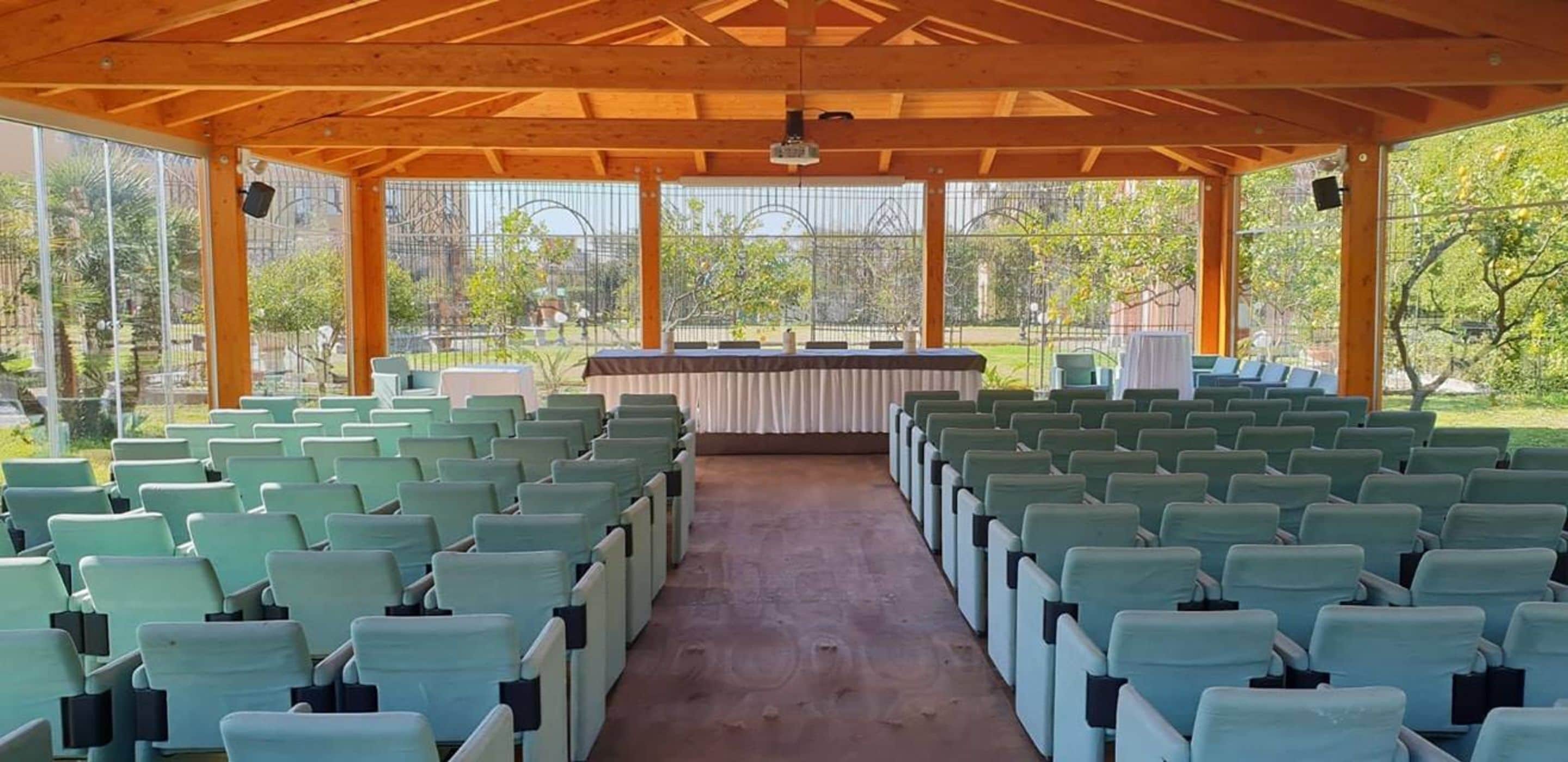 A covered outdoor seating area with rows of chairs and a stage with a table and chairs.