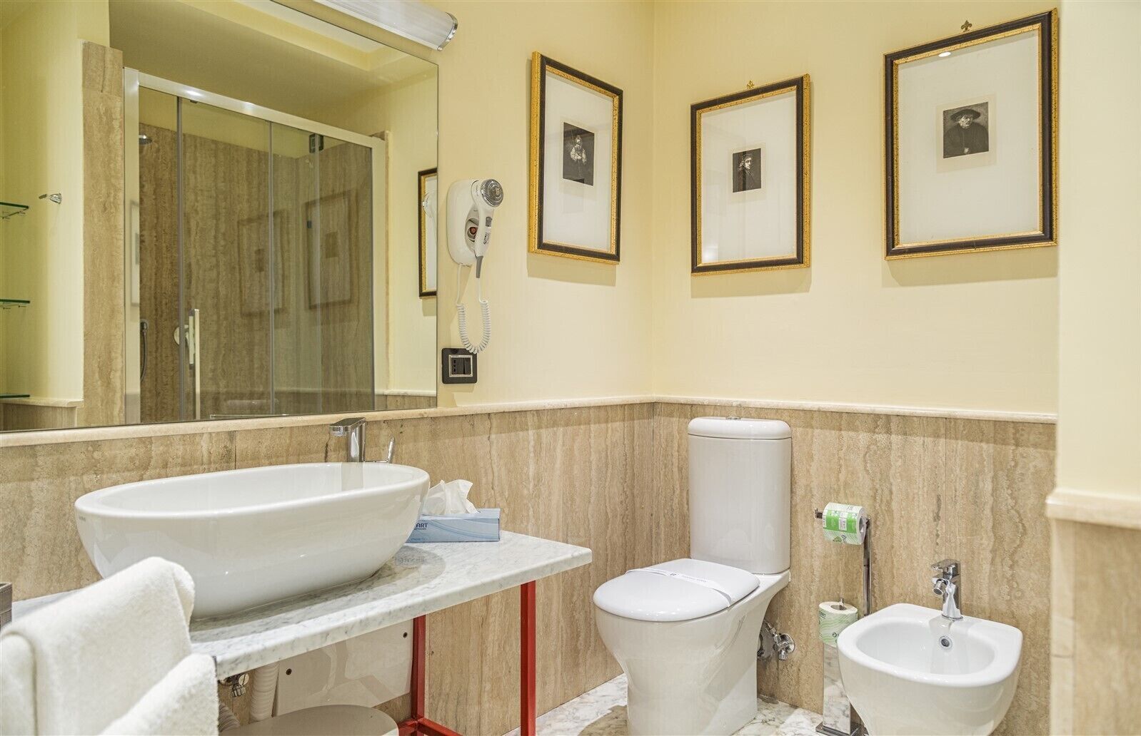 Et badeværelse med en stor vask, toilet, bidet og et spejl. Væggene er dekoreret med billeder, og der er en hårtørrer monteret.