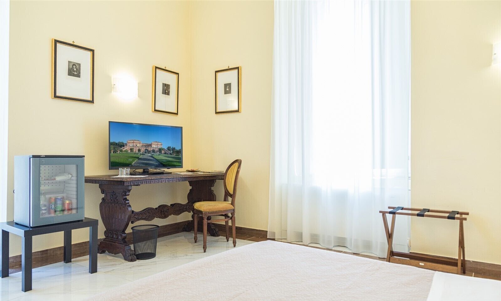 A room with a wooden desk, a chair, a television, a refrigerator, and a window with curtains.