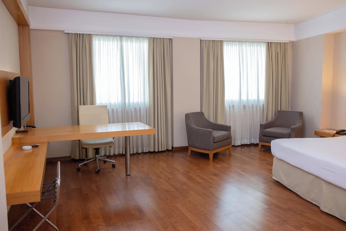 Hotellihuone, jossa on puinen työpöytä, kaksi nojatuolia, sänky ja televisio.