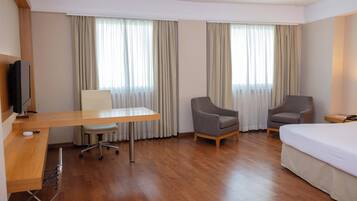 A hotel room with a wooden desk, two armchairs, a bed, and a television.