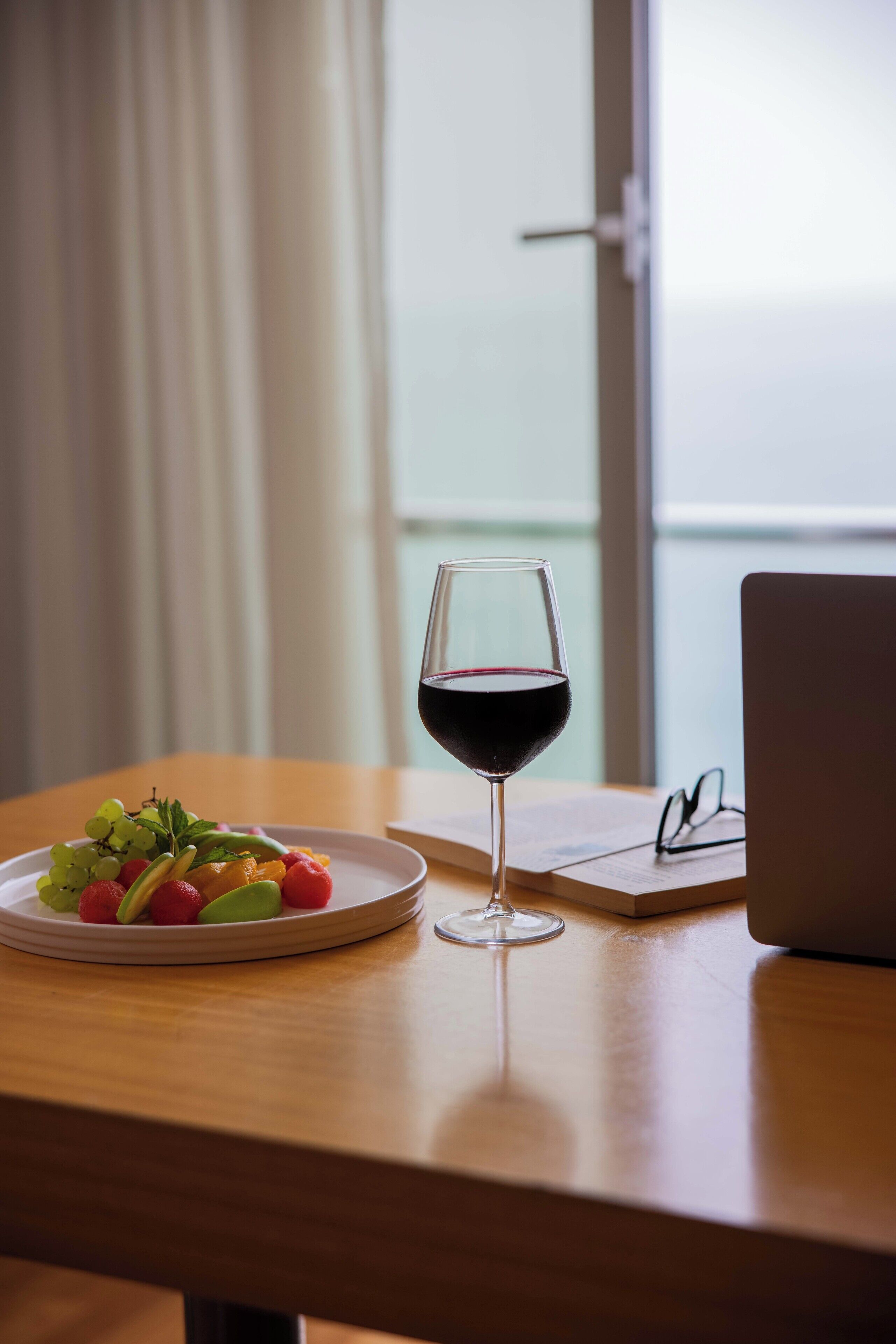 A wooden table with a plate of fruit, a glass of red wine, and a laptop.