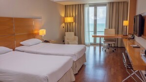 Quarto de hotel com duas camas, uma escrivaninha, uma cadeira, uma TV e vista para o mar.