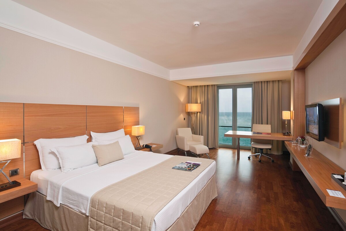 Habitación de hotel con cama, mesitas de noche, escritorio, silla, televisión y vistas al mar.