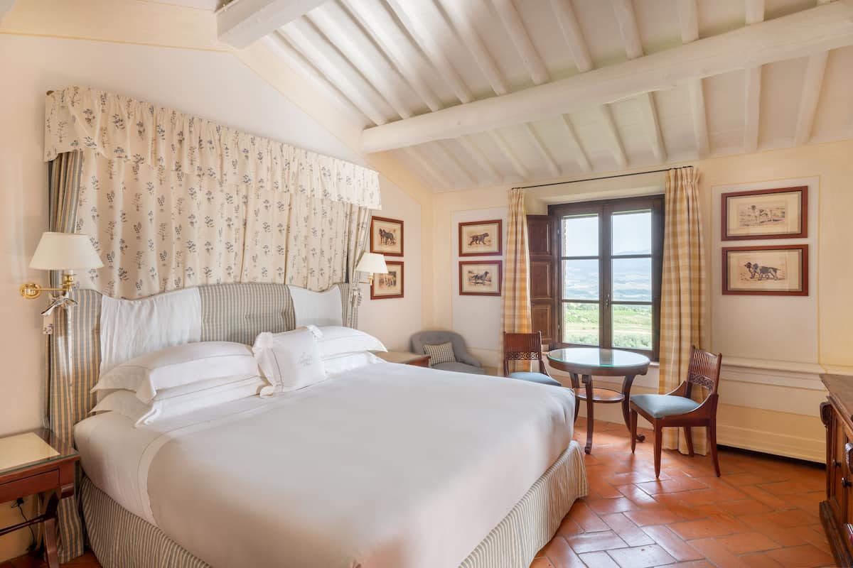 A hotel room with a large bed, a desk with a lamp, a chair, a small table, and a window with curtains.