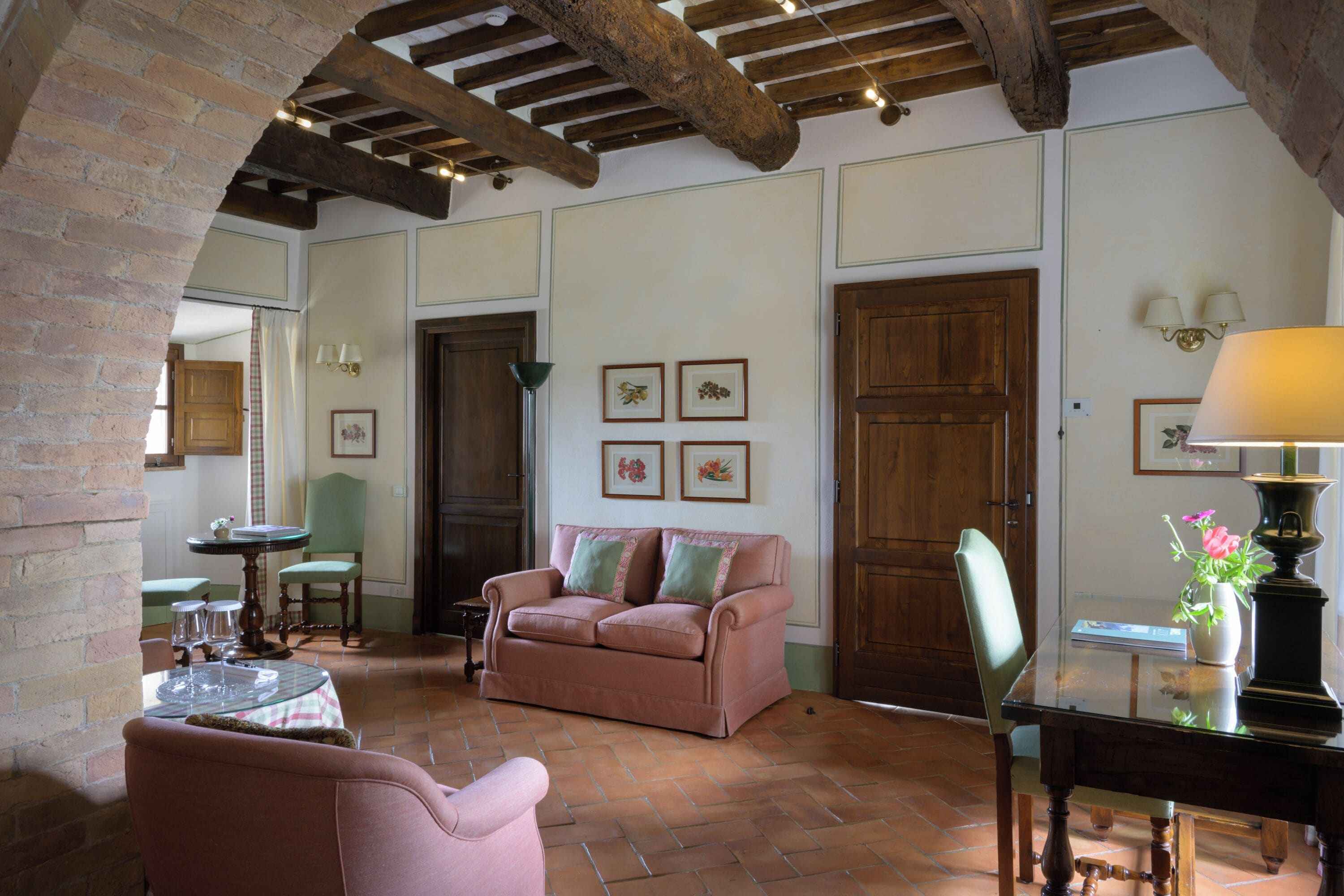 A cozy living room with a wooden ceiling, a pink sofa, a dining table with chairs, and a vase with flowers.