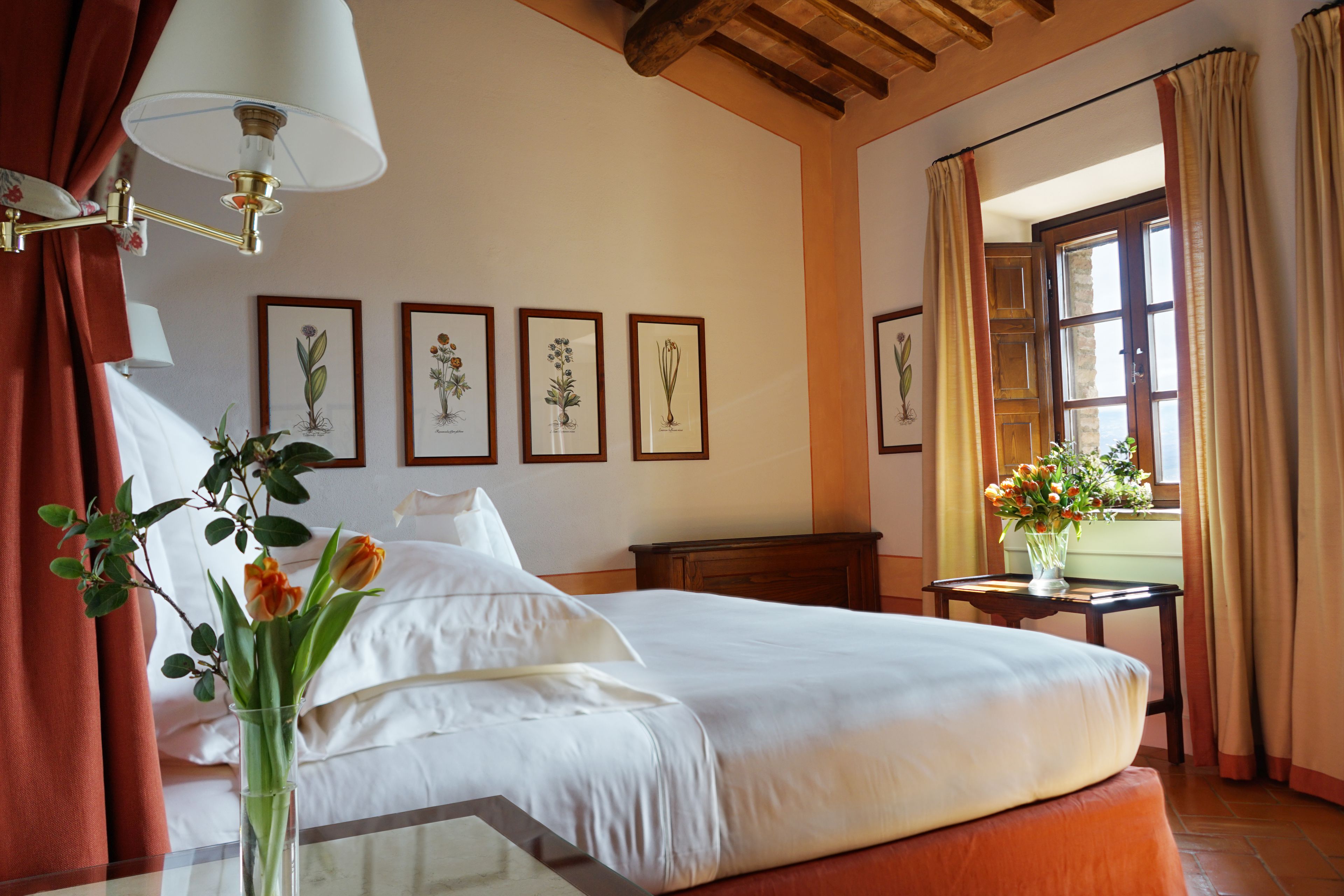 Habitación de hotel con cama, mesita de noche con un florero, lámpara e impresiones botánicas enmarcadas en la pared.