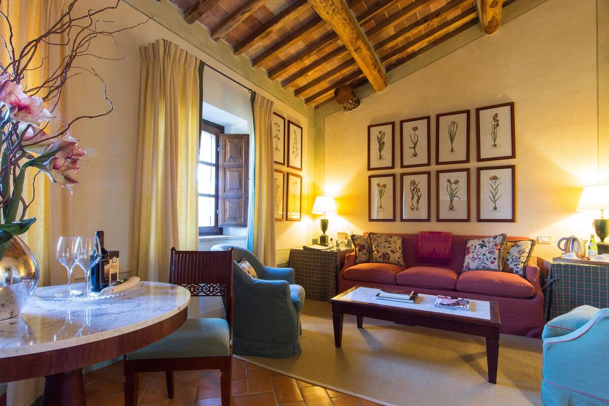 A cozy living room with a wooden ceiling, a round marble table with wine glasses, a blue armchair, a red sofa, and framed botanical artwork on the wall.