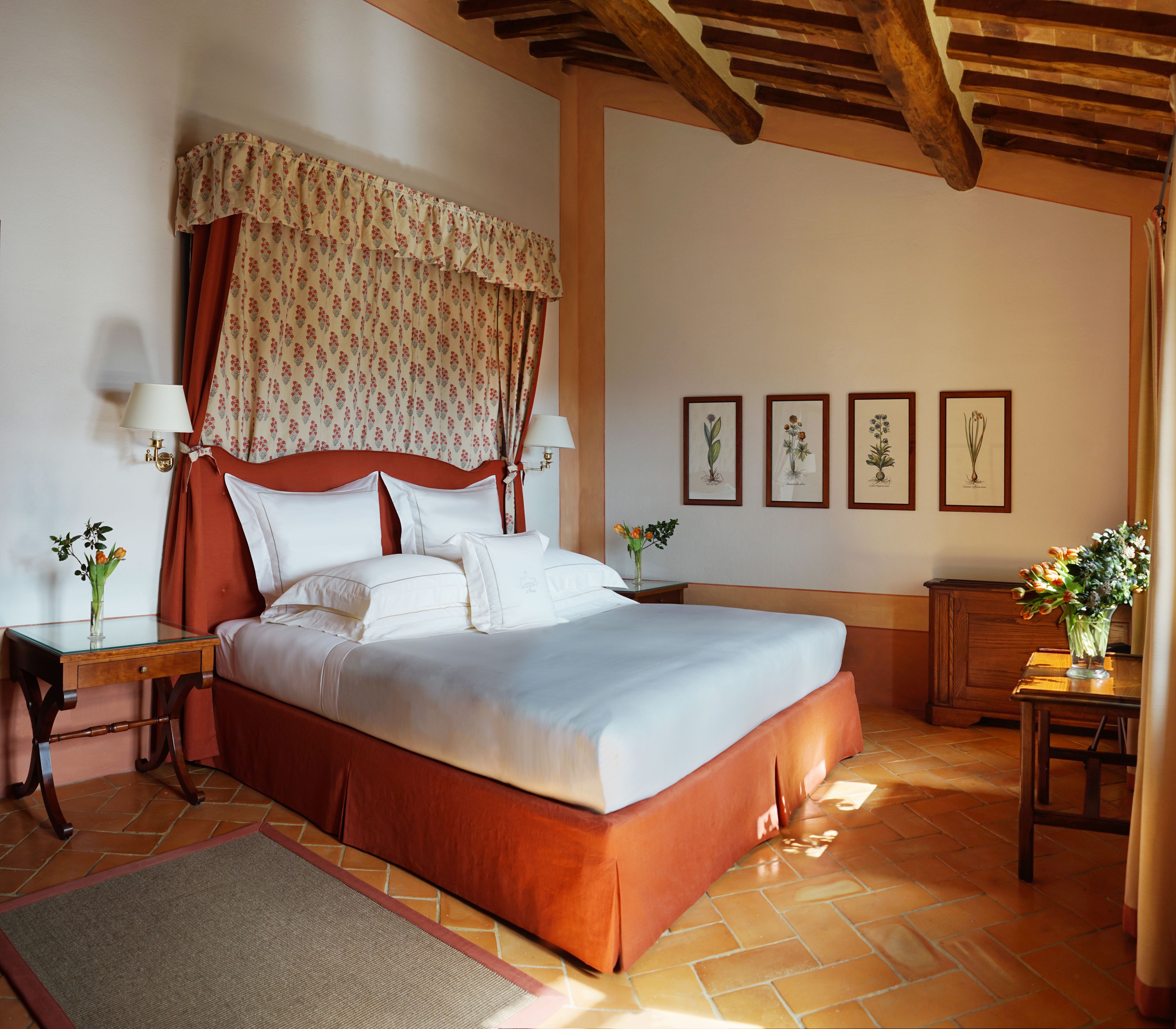 Un dormitorio amplio con una cama grande, cabecera de madera y suelo de baldosas. Hay dos mesitas de noche con lámparas y una pequeña mesa con un florero.