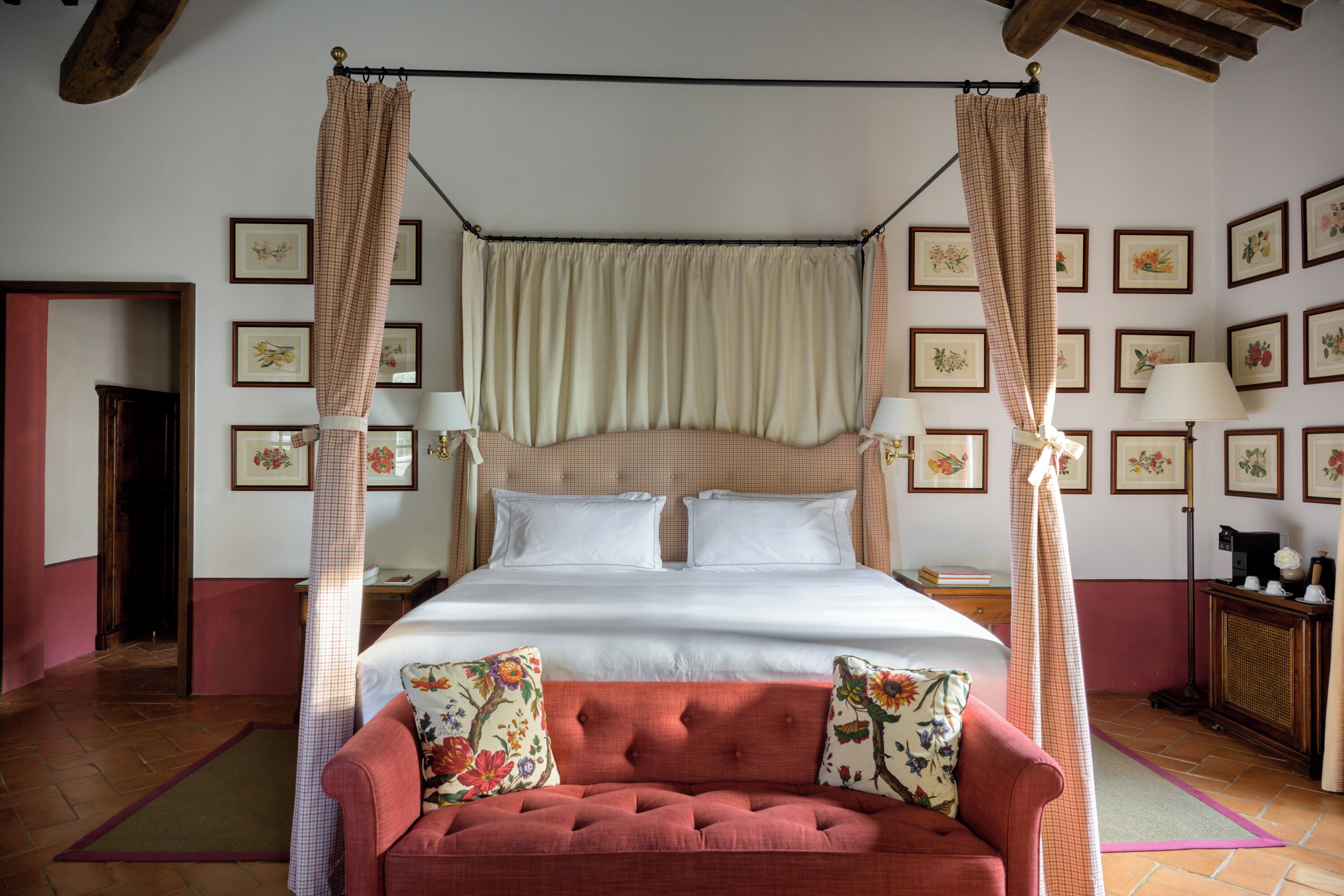 A four-poster bed with a canopy, a red sofa with floral cushions, and framed floral artwork on the walls.