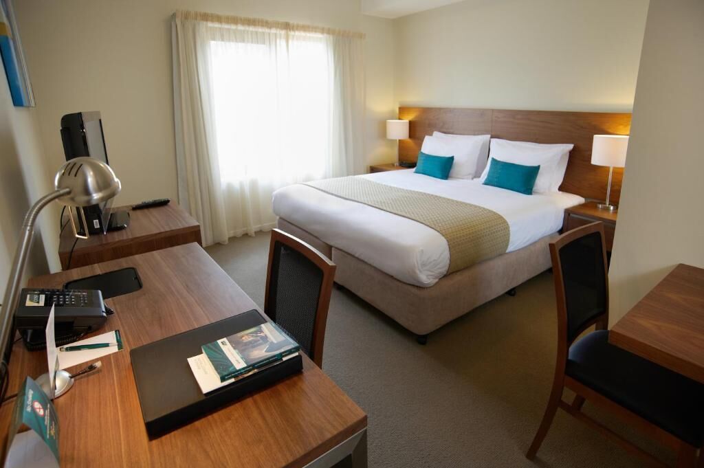 Habitación de hotel con cama, escritorio con computadora, silla, lámpara y ventana con cortinas.
