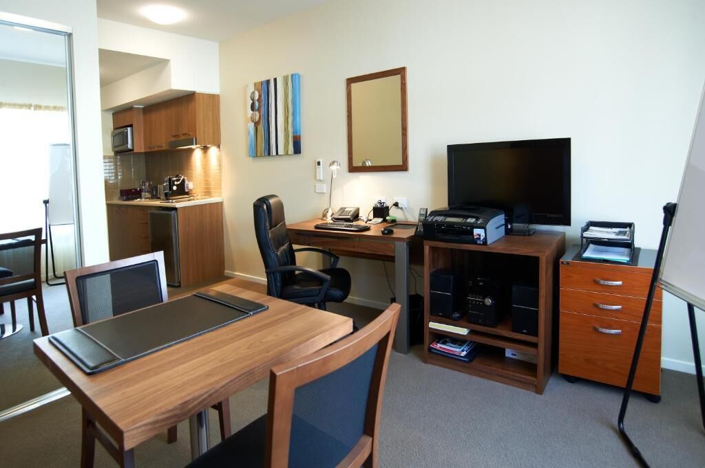 A hotel room with a wooden desk, a computer, a flat-screen TV, a kitchenette, and a dining area.