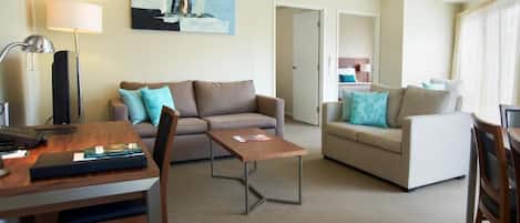 A modern living room with a brown sofa, a white sofa, and a wooden coffee table. There is a desk with a lamp and a laptop.