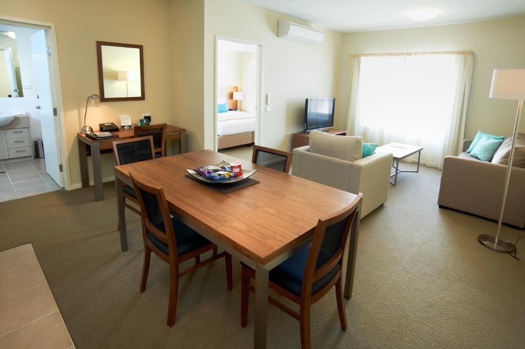A hotel room with a wooden dining table, two chairs, a sofa, a TV, and a lamp.