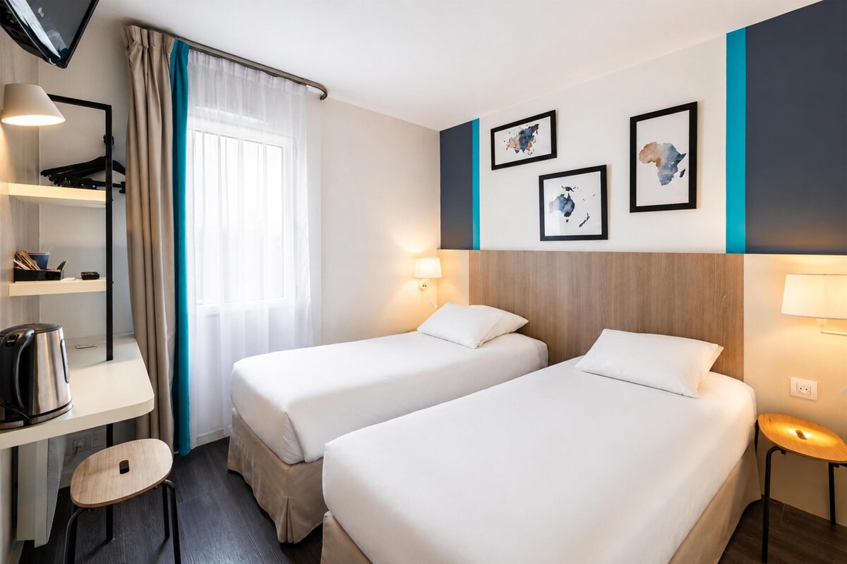 Habitación de hotel con dos camas, un escritorio, una tetera, una silla y cuadros en la pared.