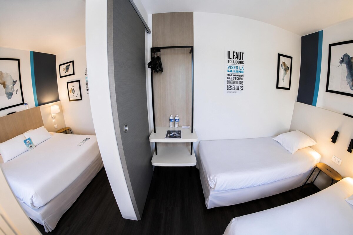 Habitación de hotel con dos camas, una mesita y una botella de agua, además de una pared con una frase motivacional.