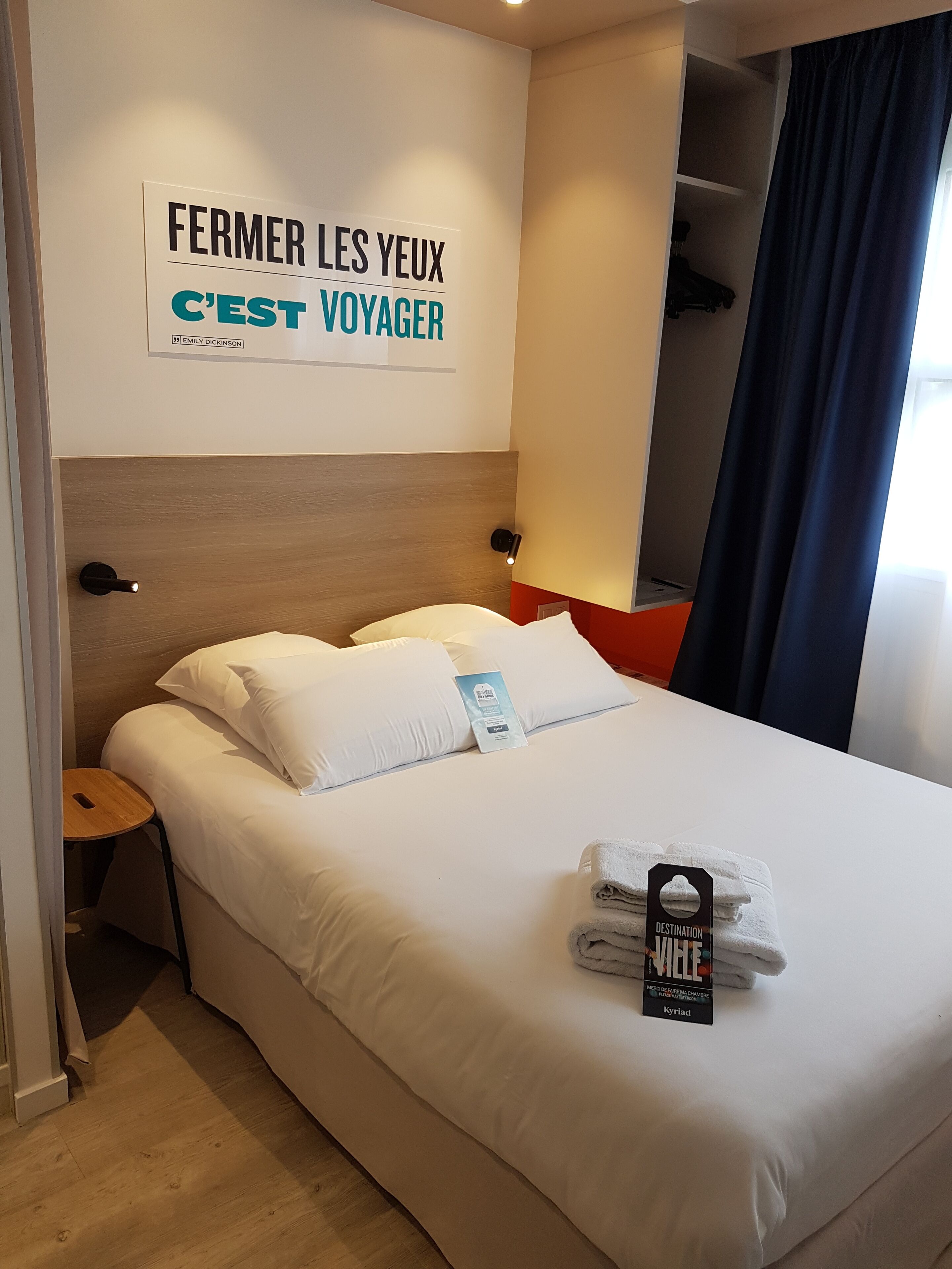 Una habitación de hotel con cama, mesita de noche, una pared con un cartel, ventana con cortinas y un armario.