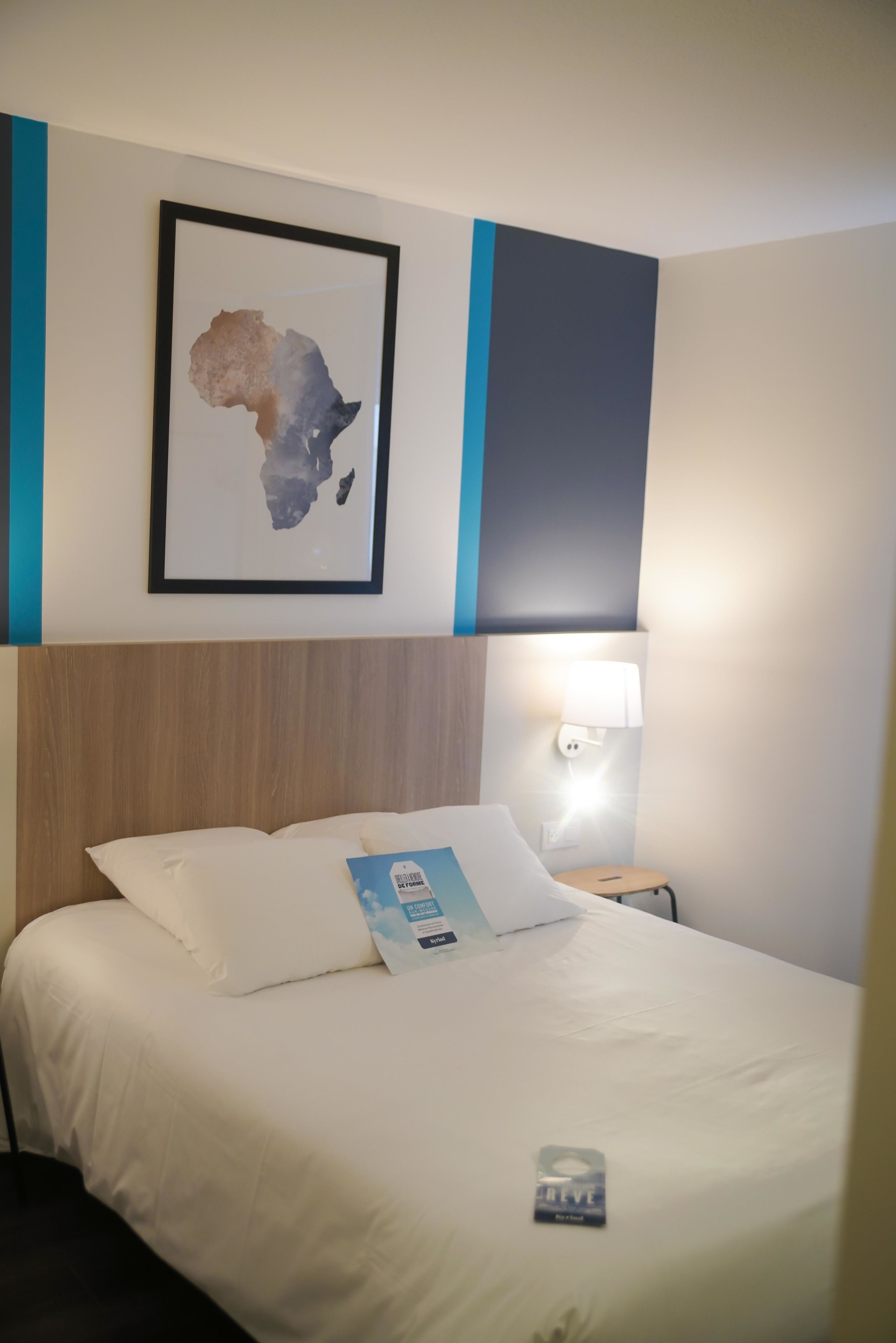 Ein Hotelzimmer mit einem Bett, einer gerahmten Karte an der Wand, einem Nachttisch mit Lampe und einer Zeitschrift auf dem Bett.
