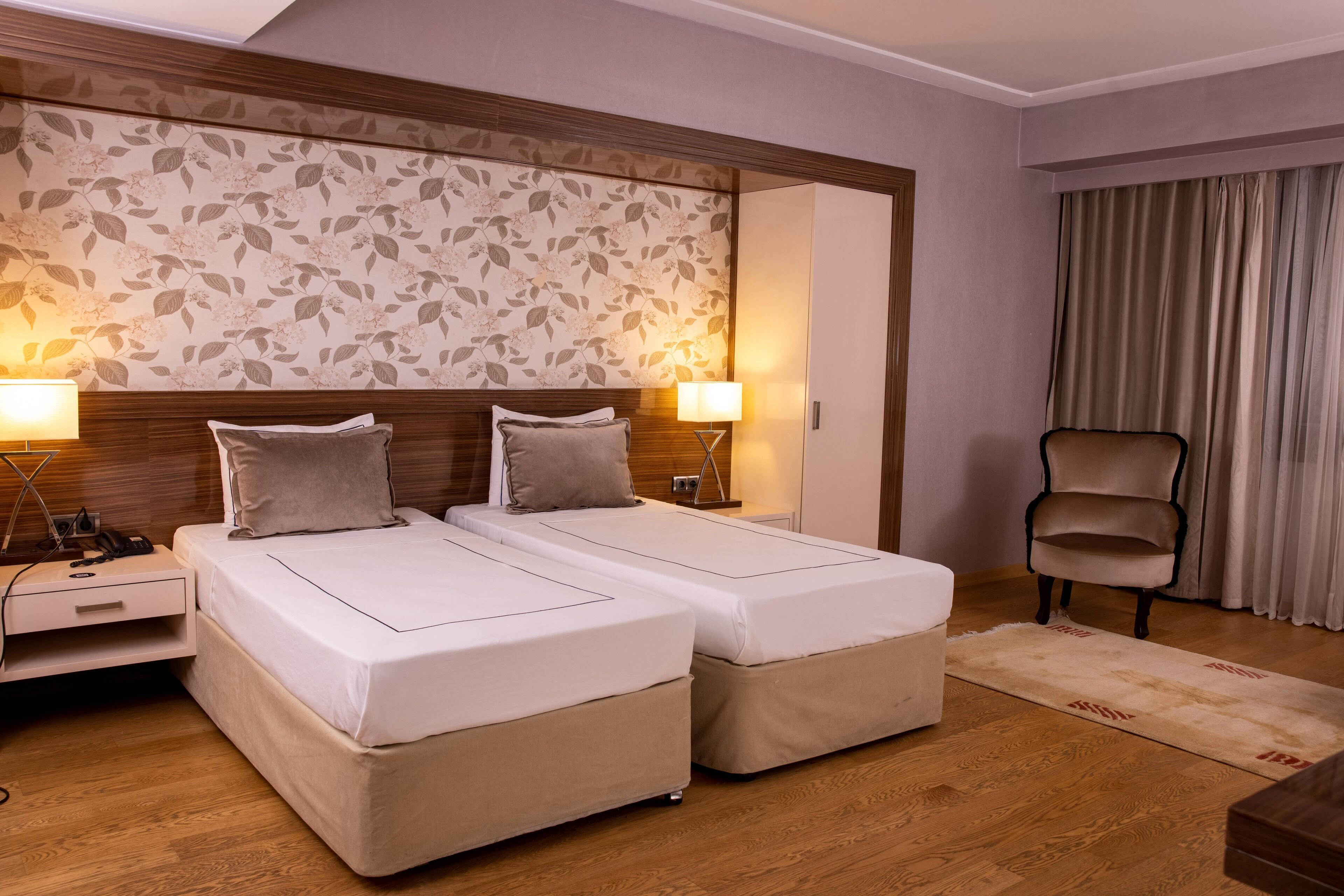 Een hotelkamer met twee bedden, een houten hoofdeinde, bloemen behang, twee bedlampjes, een stoel en een houten vloer.