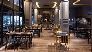 Een modern restaurant met houten tafels en stoelen, een bar en uitzicht op de stad door grote ramen.
