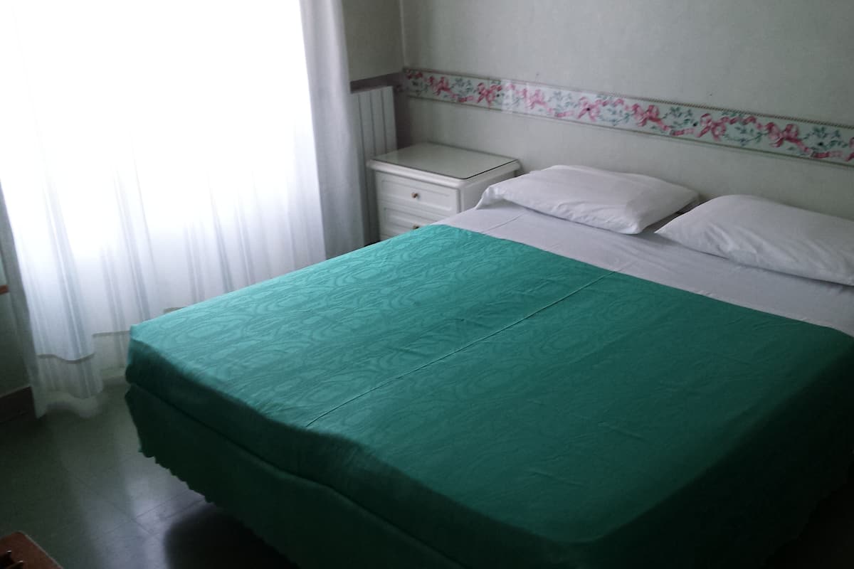 A single-bed room with a green bedspread, white pillows, a nightstand, and a dresser.
