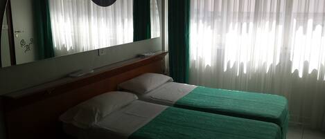 Habitación de hotel con dos camas individuales, cabecero de madera, un espejo grande y cortinas de gasa.