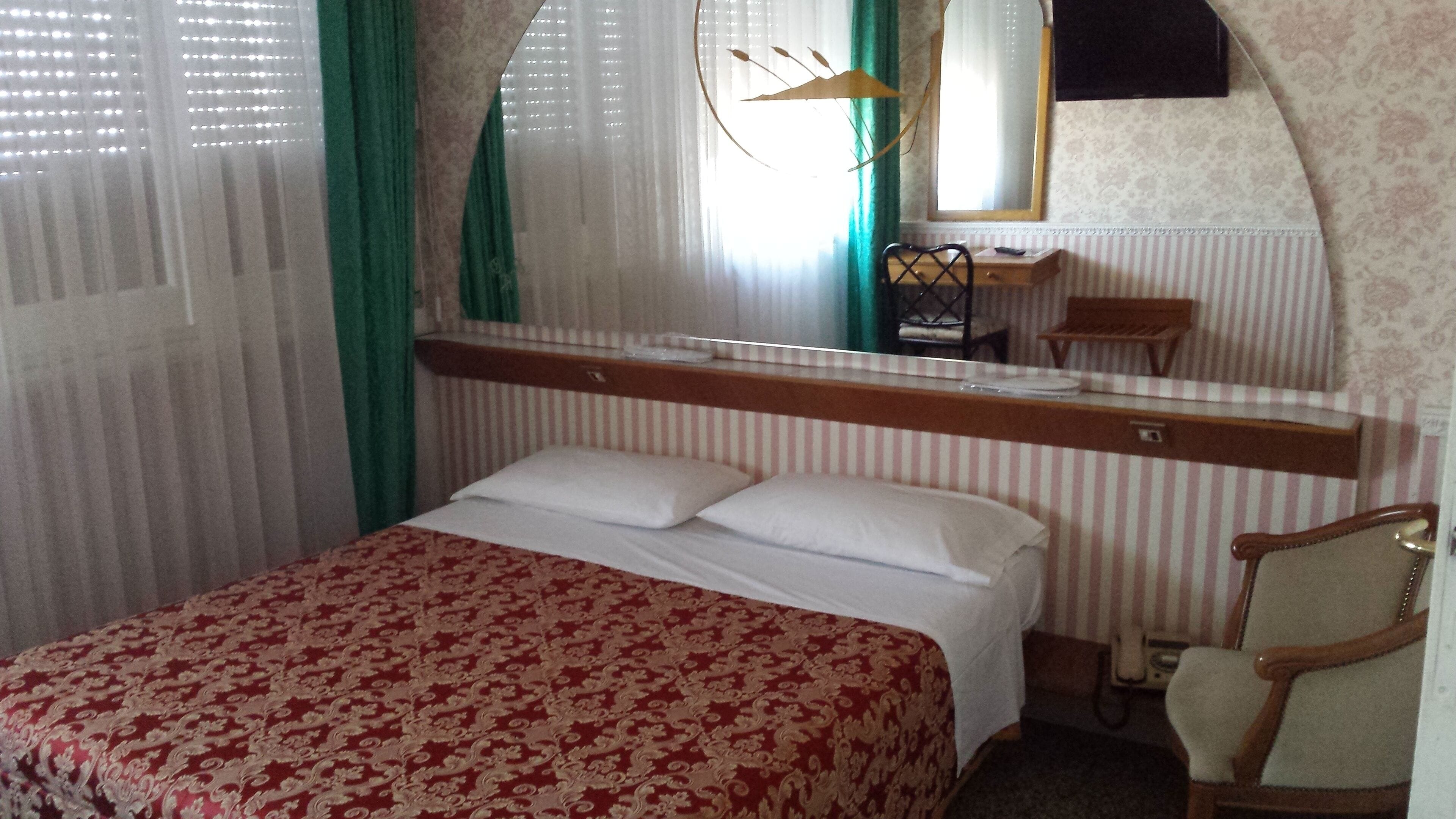 Una habitación de hotel con una cama, una silla, una mesita de noche, un espejo y una ventana con cortinas.