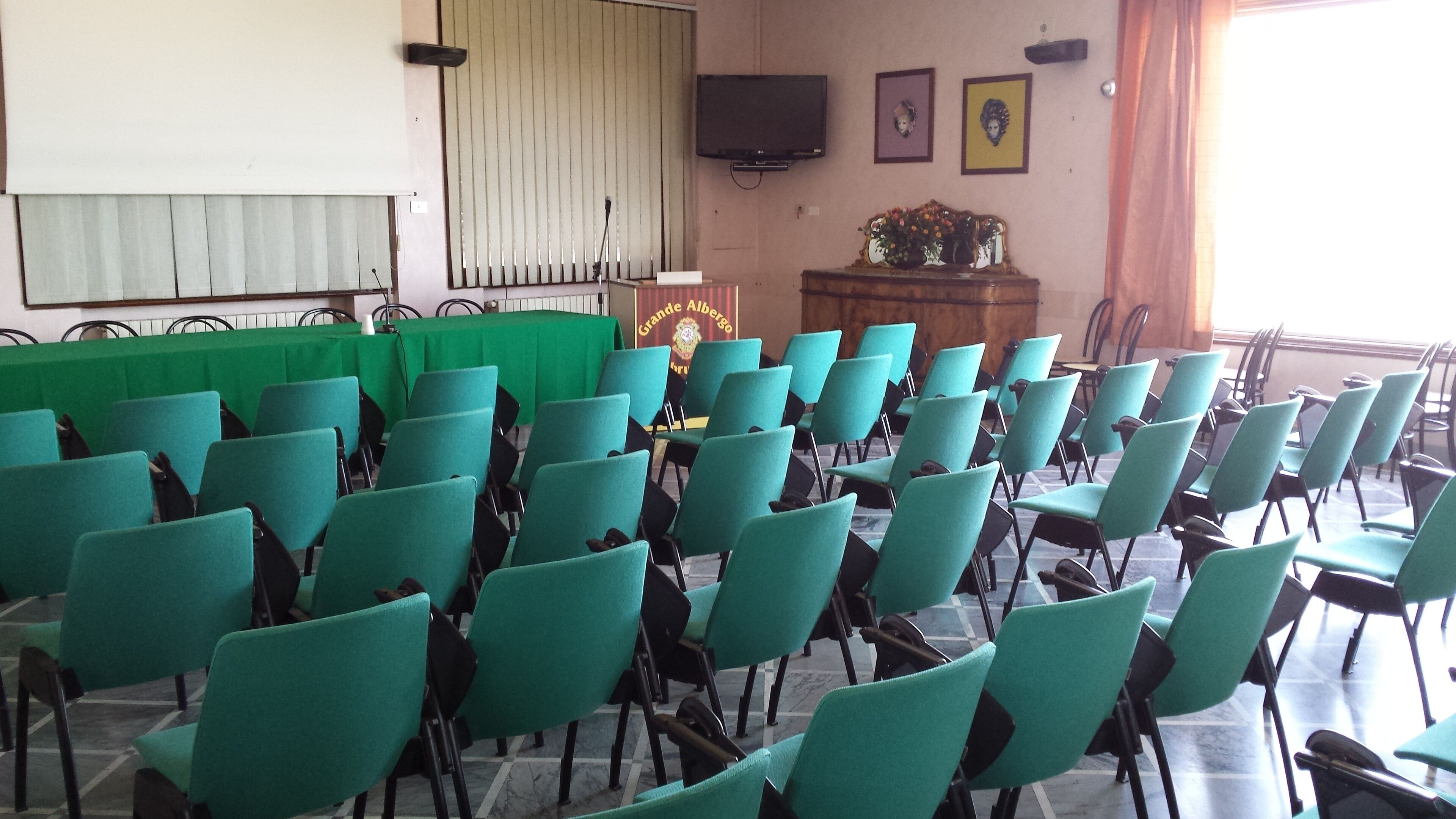 A conference room with rows of green chairs, a podium, a television on the wall, and a banner with a crest.