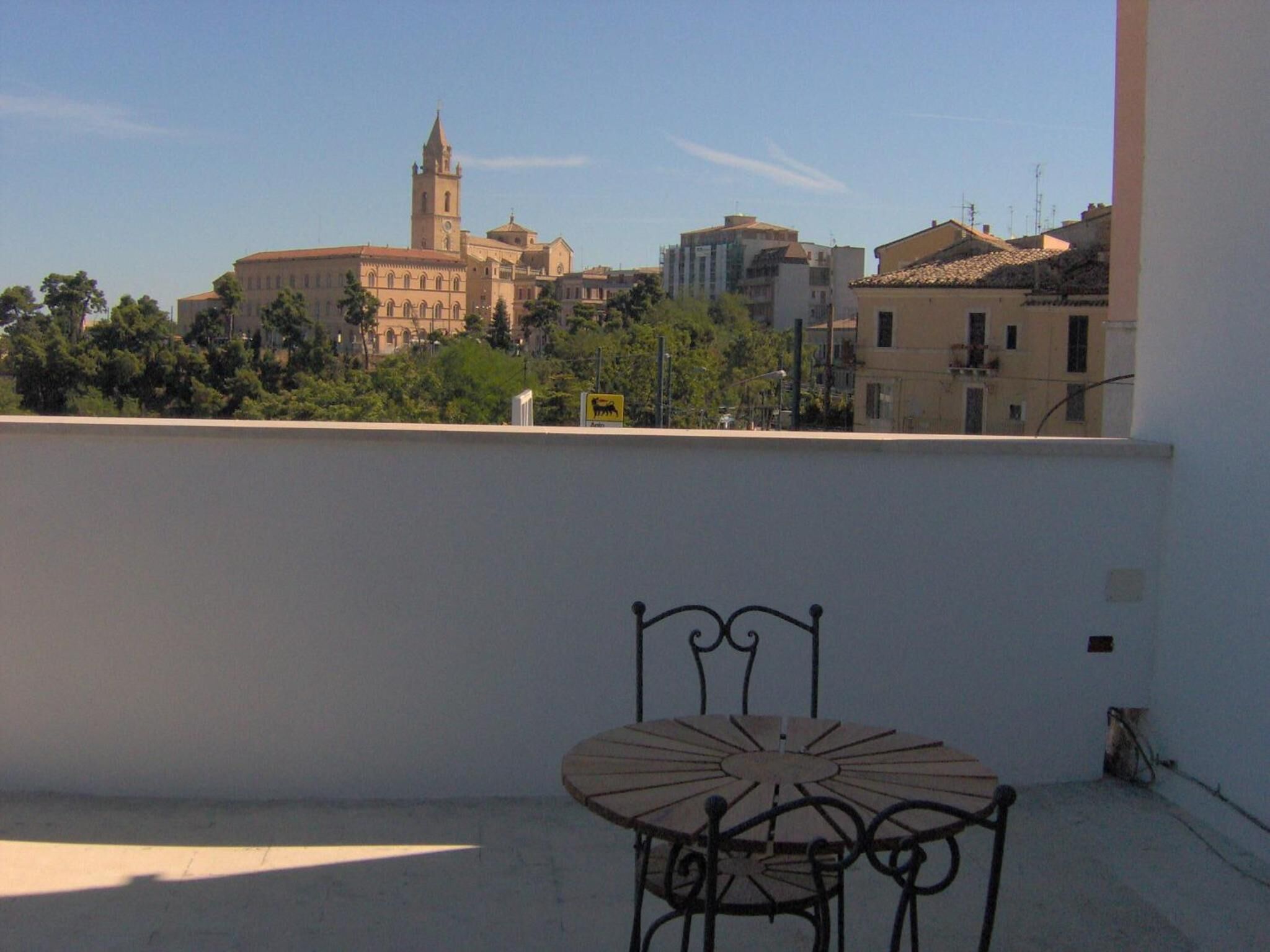 Un balcón con una silla y una mesa de metal, con vistas a un paisaje urbano que incluye edificios históricos y una torre de iglesia.