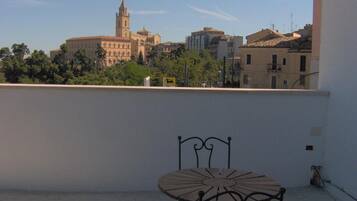 Un balcón con una silla y una mesa de metal, con vistas a un paisaje urbano que incluye edificios históricos y una torre de iglesia.