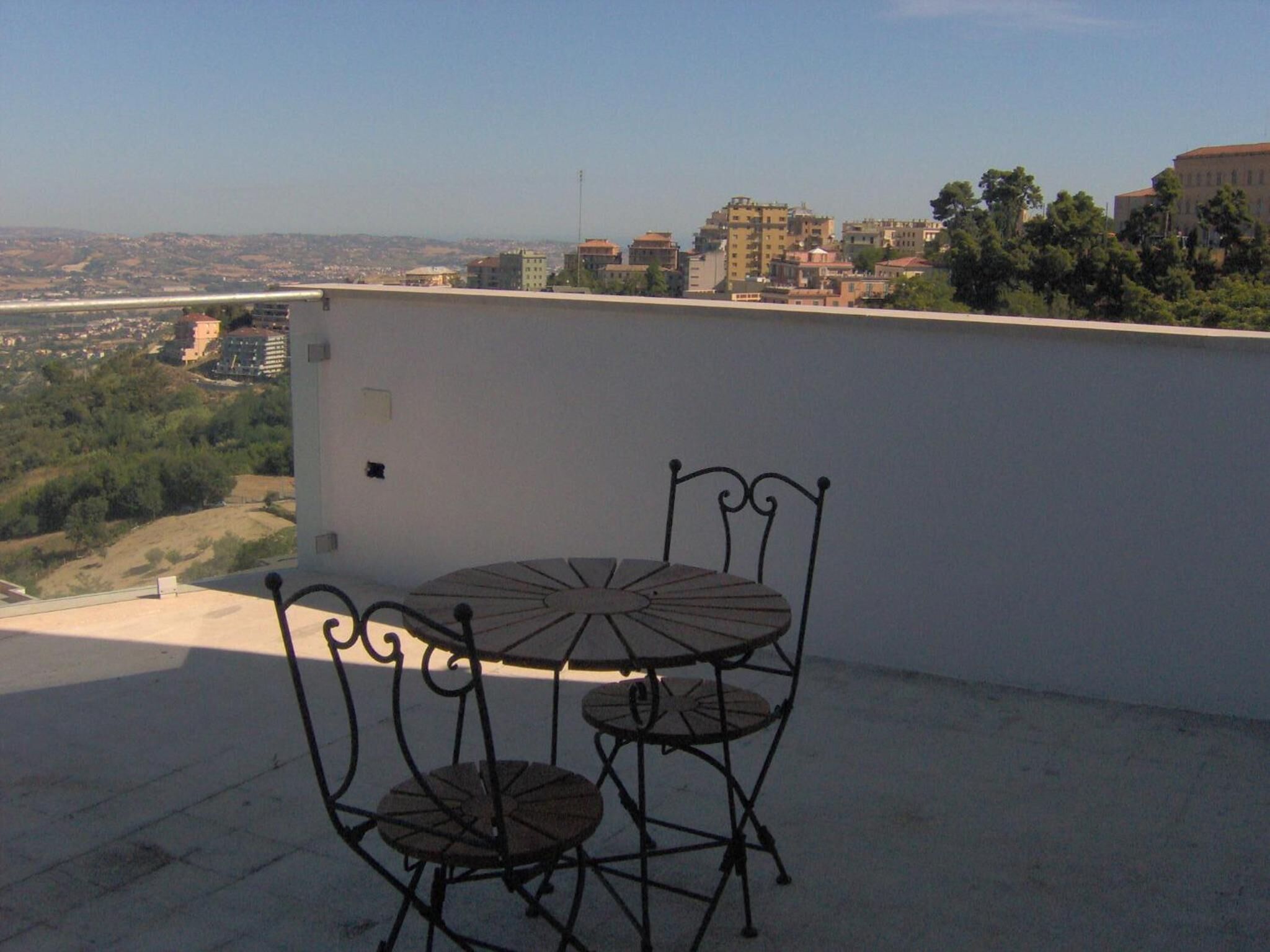 Una terraza en la azotea con una mesa y sillas de metal, con vistas a un paisaje urbano con edificios y zonas verdes.