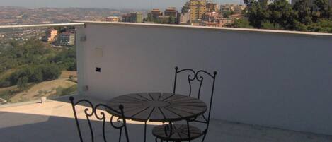 Una terraza en la azotea con una mesa y sillas de metal, con vistas a un paisaje urbano con edificios y zonas verdes.