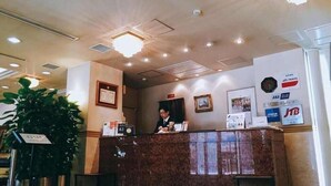 A hotel reception desk with a receptionist, a clock, and various signs including ANA and JTB. - Nissin Namba Inn (Osaka)