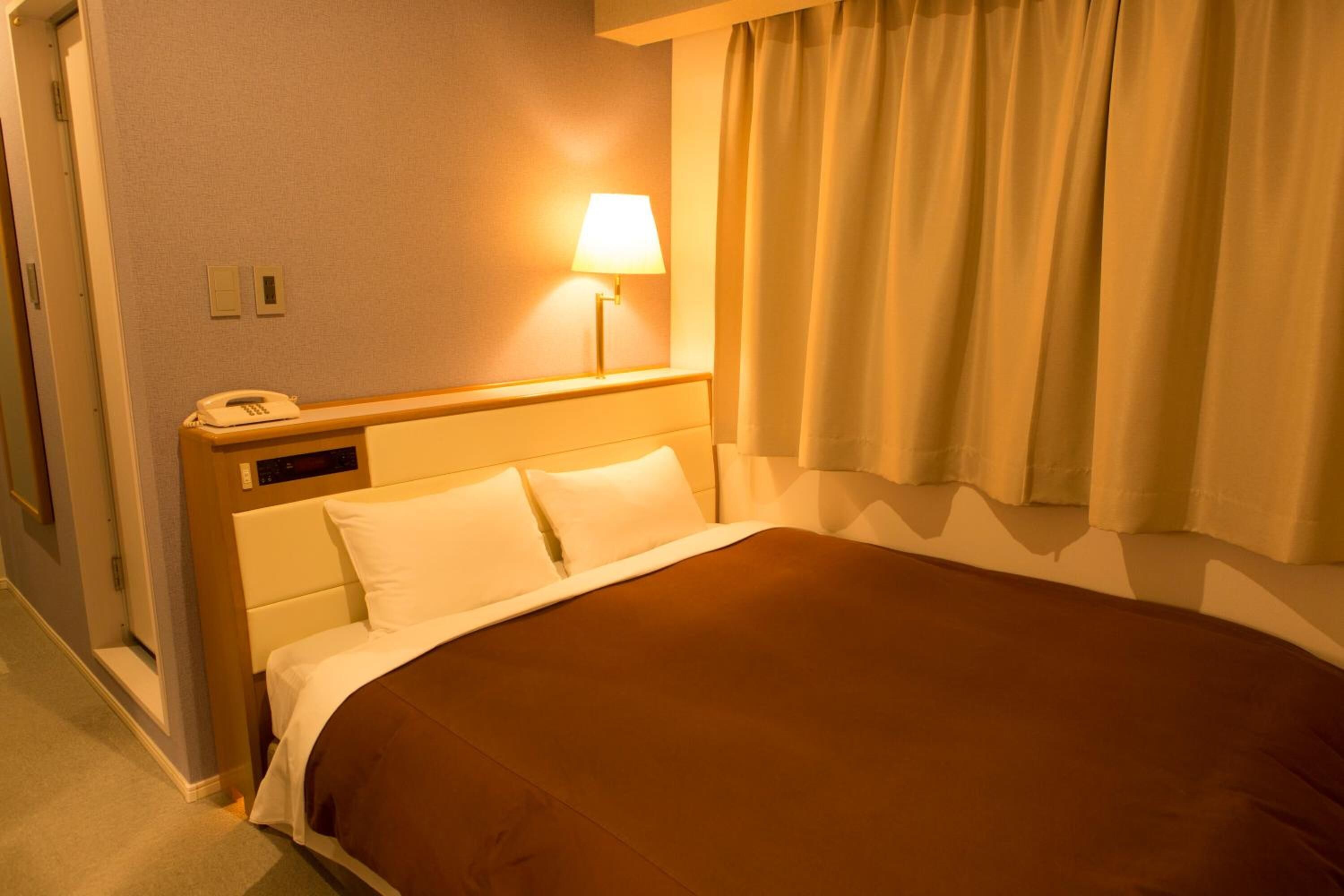A hotel room with a bed, a bedside lamp, a telephone, and a window with curtains.