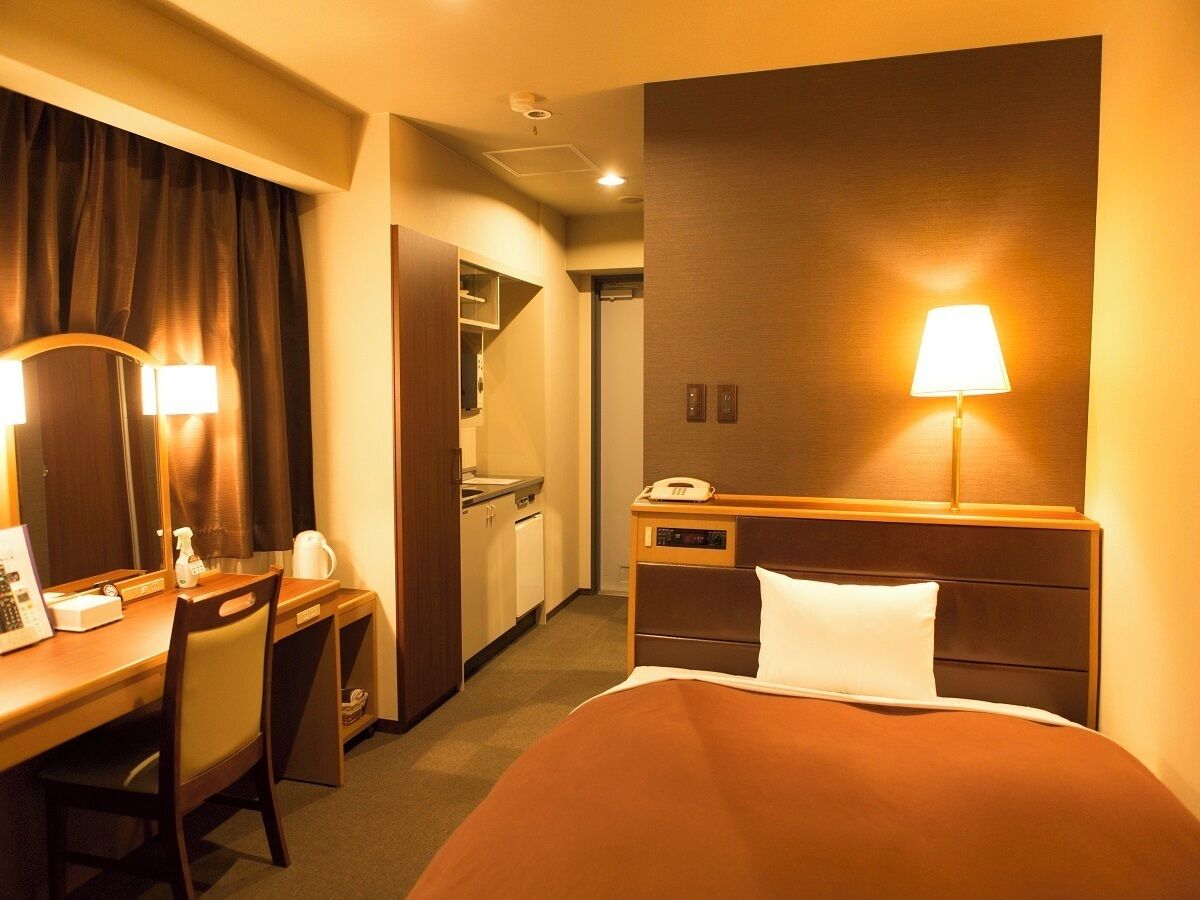 A hotel room with a bed, desk, chair, lamp, and a kitchenette.