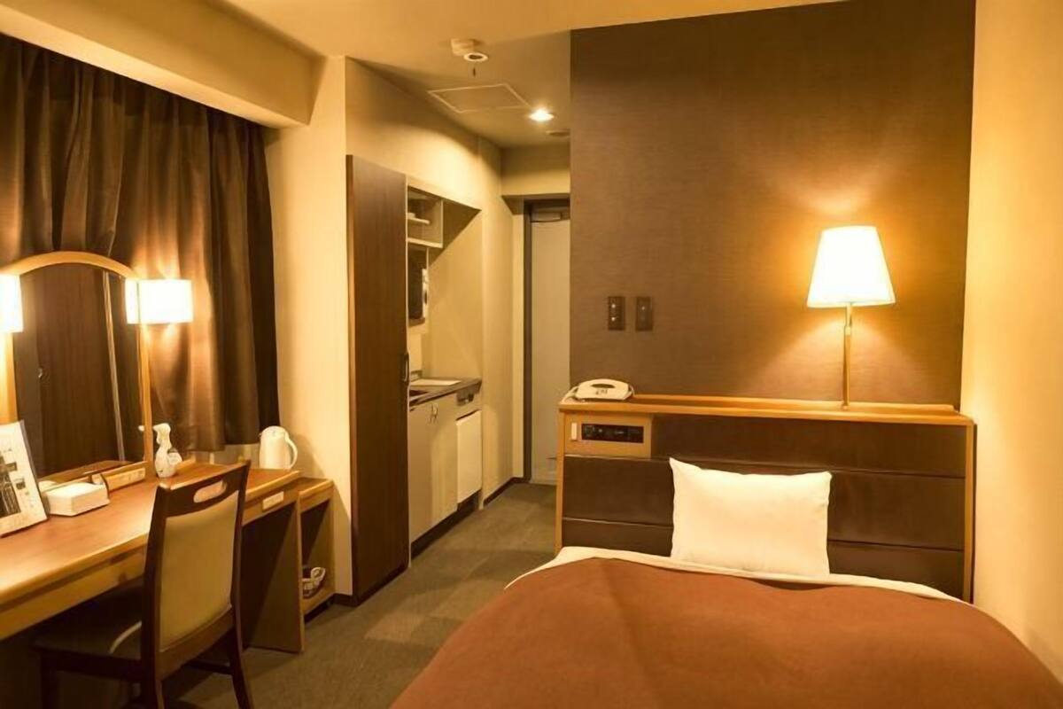A hotel room with a bed, desk, chair, lamp, and a small kitchenette.