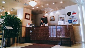 A hotel reception desk with a receptionist, a large potted plant, and various signs and posters on the walls. - Nissin Namba Inn (Osaka)