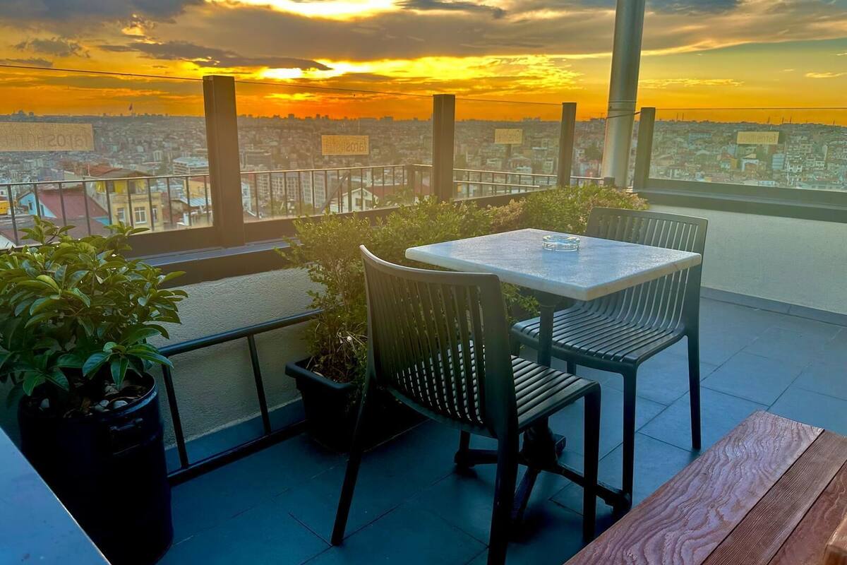 A balcony with a table and chairs, potted plants, and a view of a cityscape at sunset.