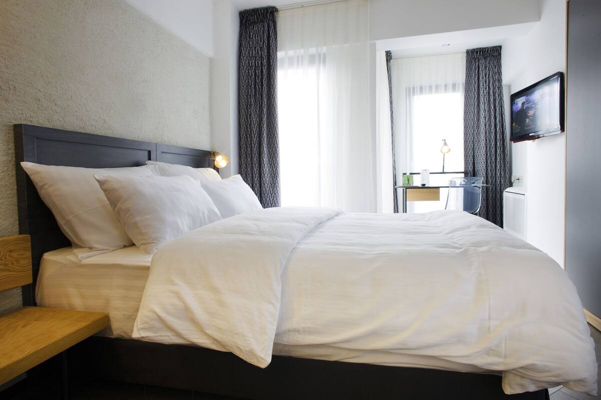 A neatly made bed with white linens and a wooden headboard, a bedside table with a lamp, and a television mounted on the wall.
