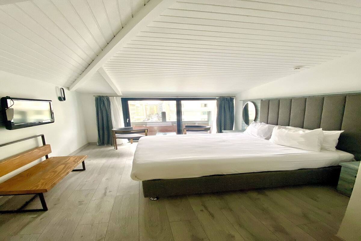 A modern hotel room with a large bed, a wooden bench, a flat-screen TV, and a view of the city through large windows.