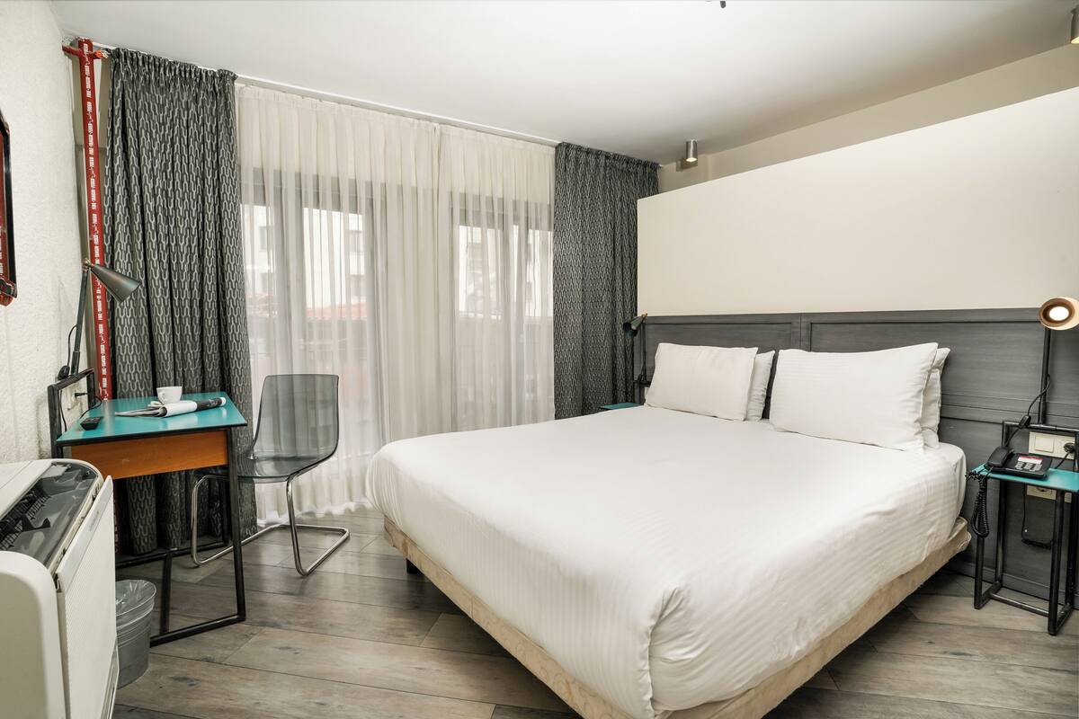 A hotel room with a large bed, a desk with a chair, a small table, and a nightstand with a lamp.