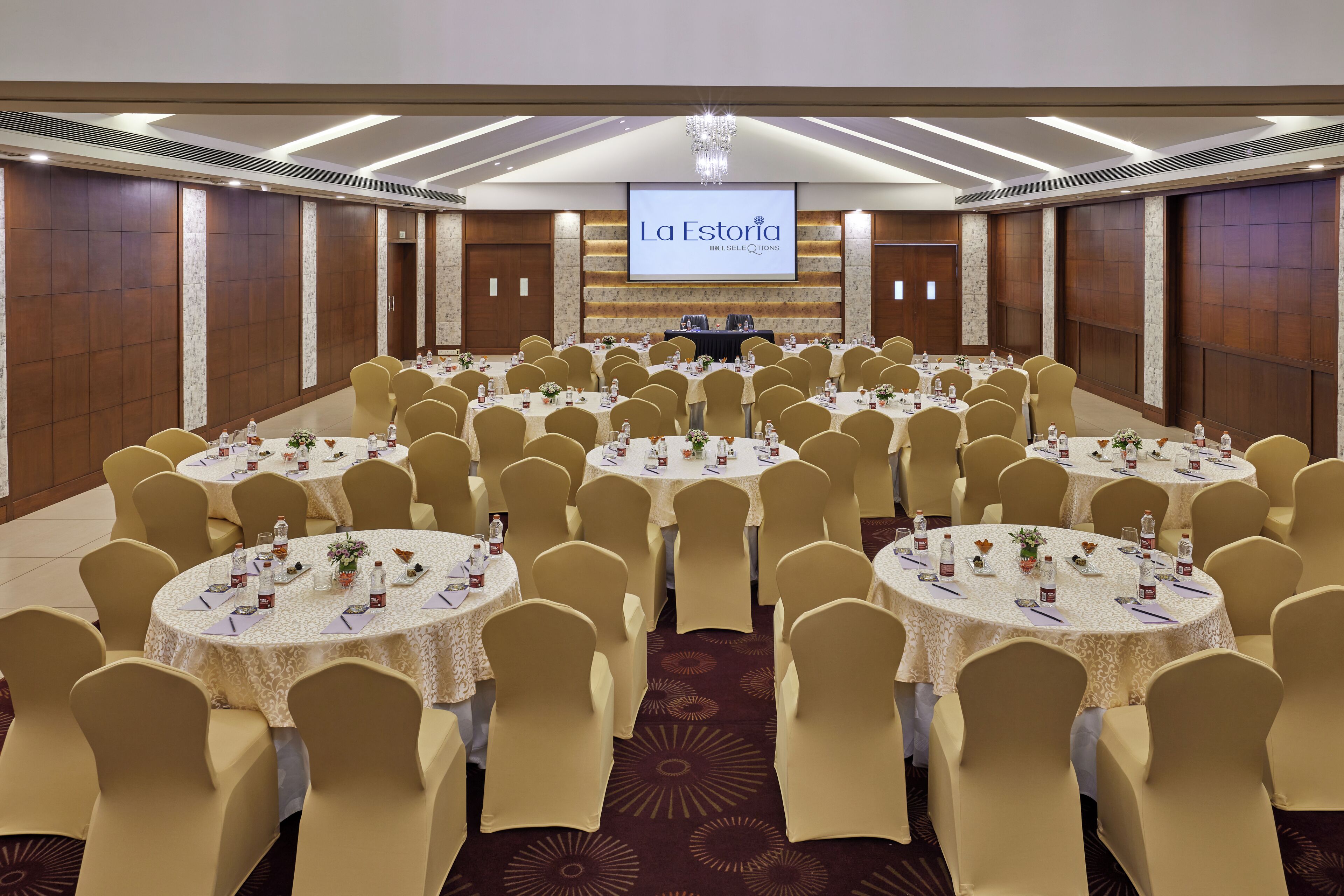 Un salón de conferencias con varias mesas redondas, sillas color beige y una pantalla grande al frente.