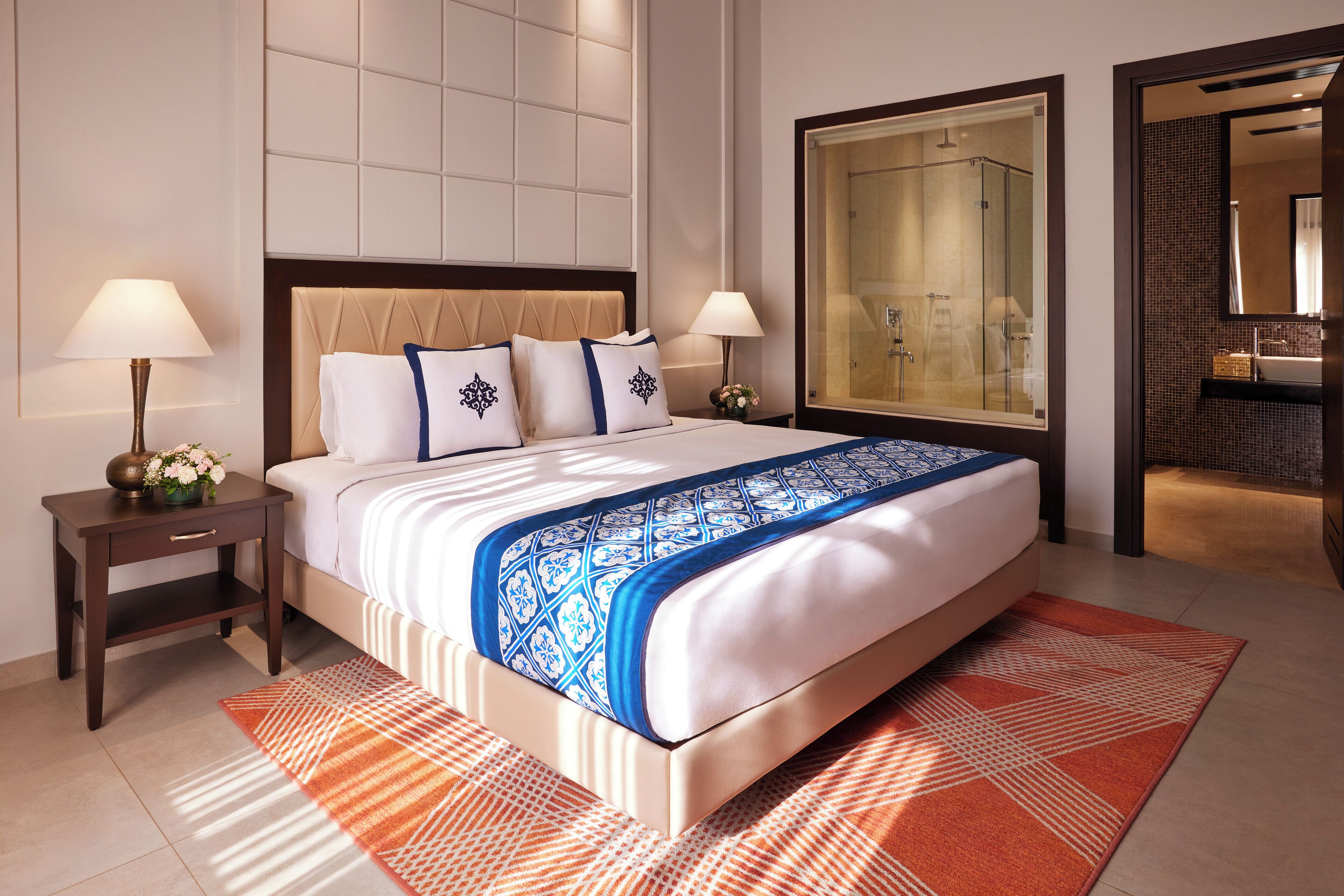 Habitación de hotel con una cama grande, dos mesitas de noche con lámparas, zona de ducha y baño con lavabo y espejo.