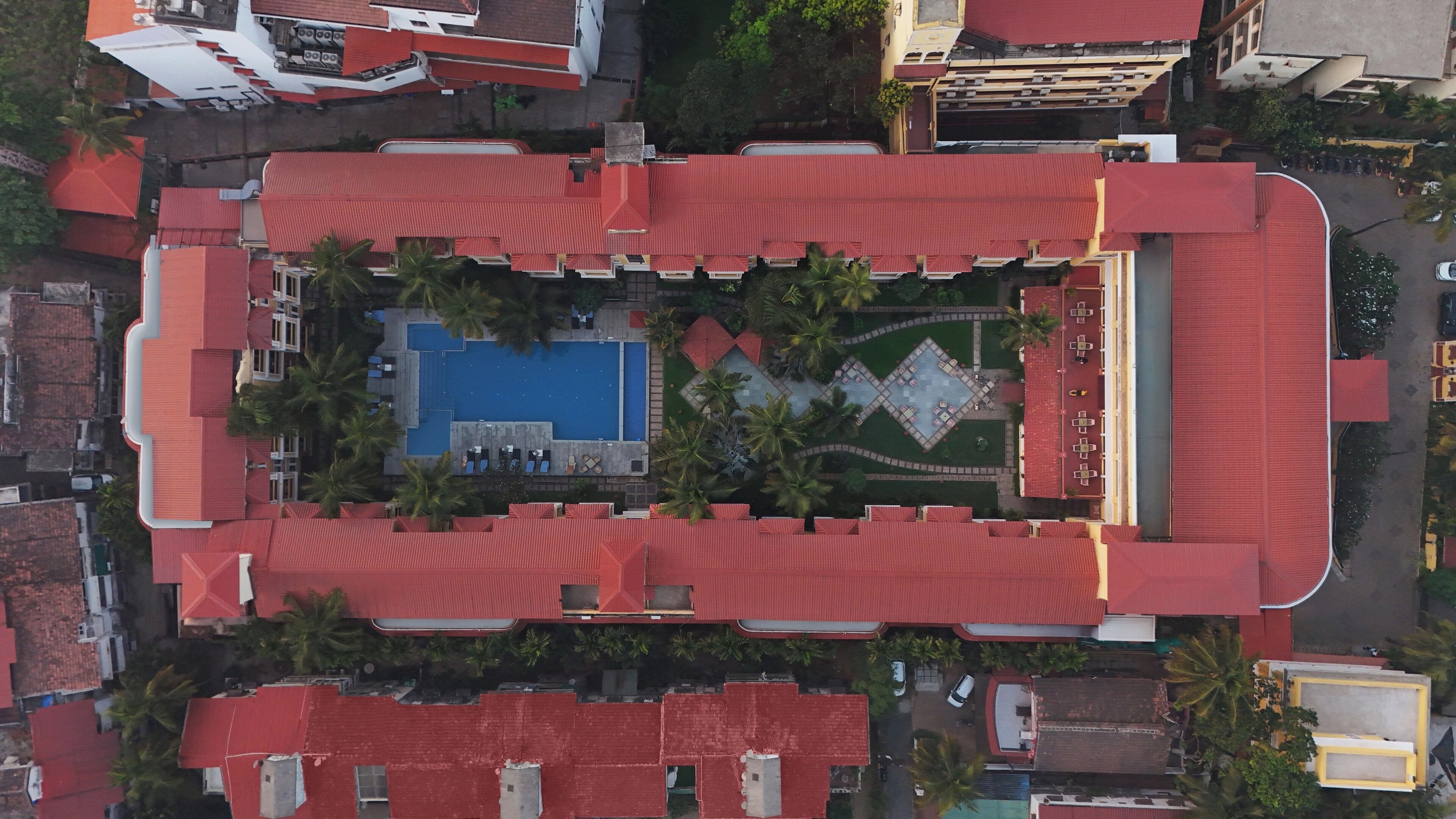 Vue aérienne d’un hôtel doté d’une piscine centrale, entourée de palmiers et d’un jardin aux formes géométriques.