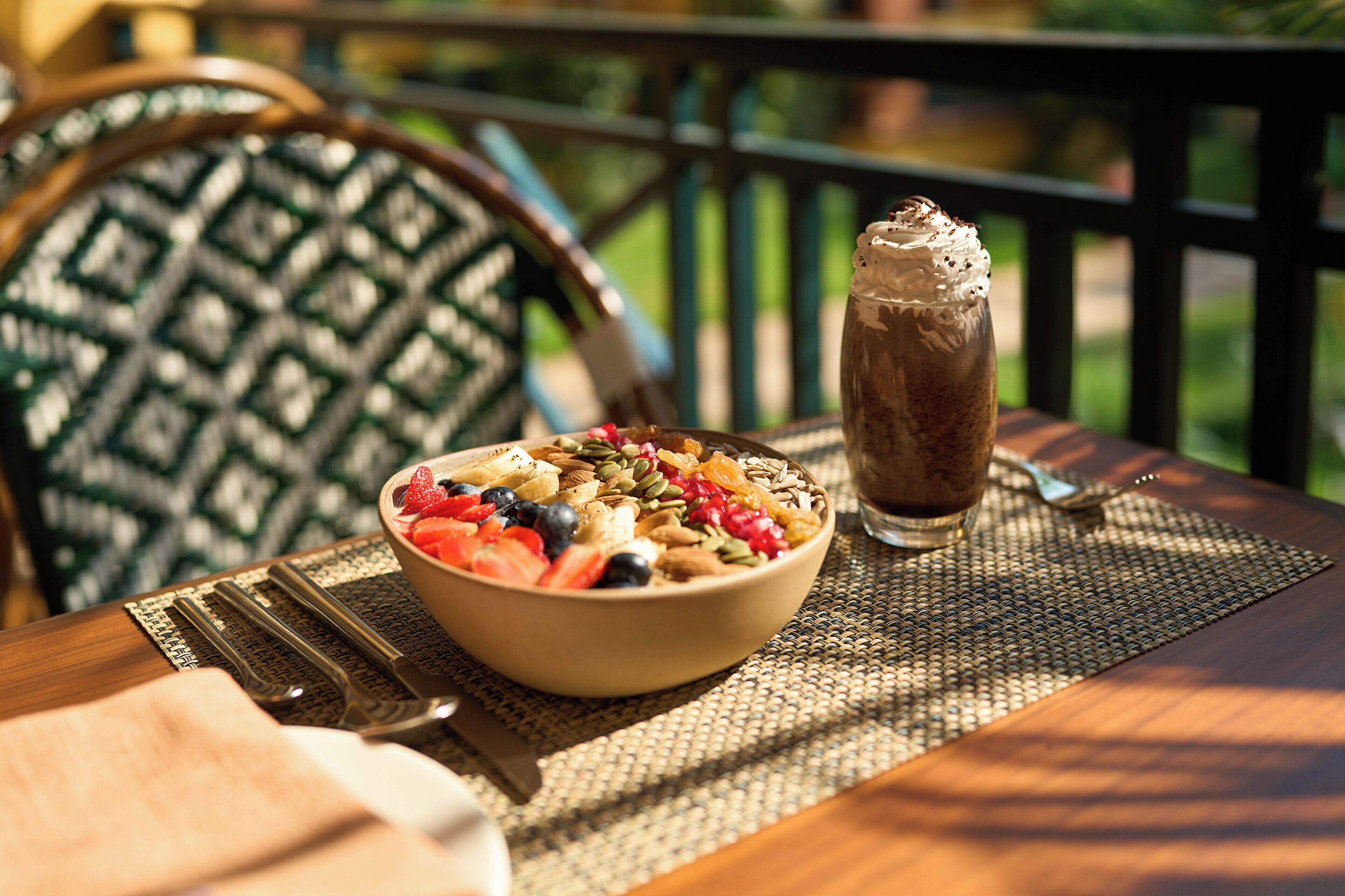 A breakfast setting with a bowl of cereal and a glass of chocolate milk on a table outdoors.